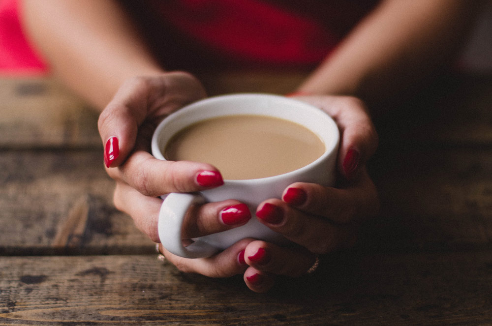 Photo of womens hands holding a cup of coffee. She has a red dress on and red nail polish and is leaning on a wooden table.